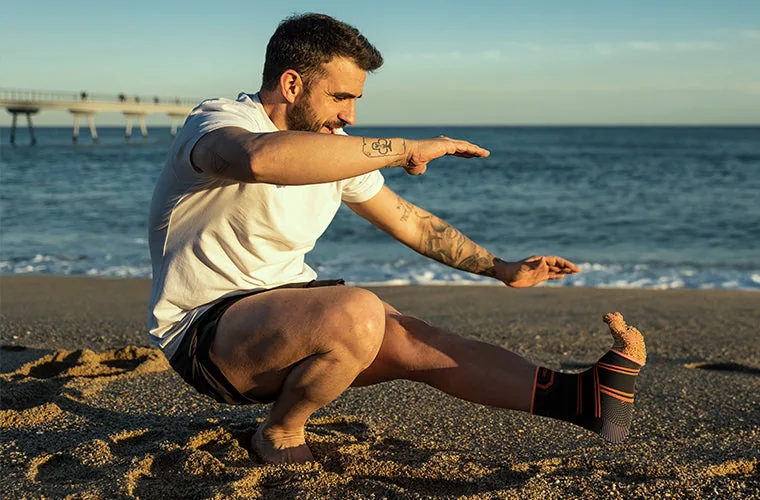 Man in a white T-shirt balancing in a one-leg squat on a beach, wearing the Tynor Ankle Binder Air Pro