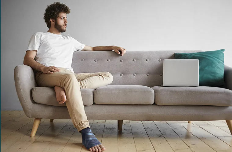 Man in a white T-shirt relaxing on a grey sofa beside a laptop, wearing the Tynor Ankle Binder
