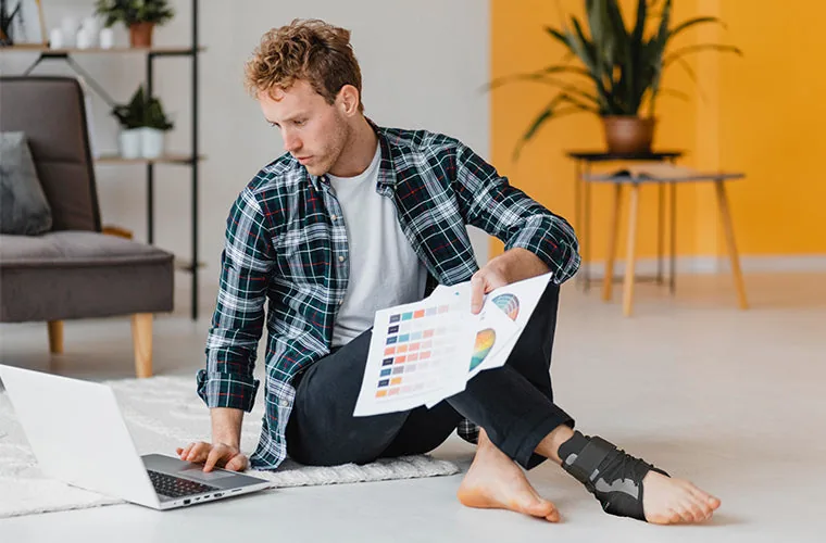 Man in a checked shirt sitting on the floor with a laptop and papers, wearing the Tynor Ankle Brace