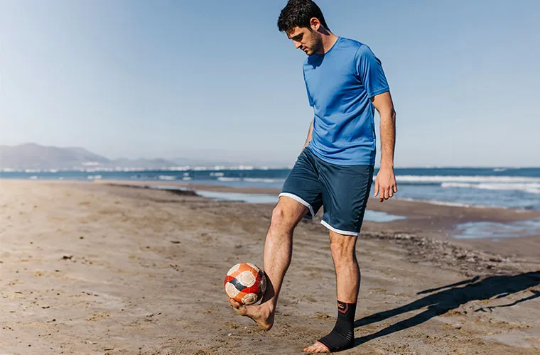 Man in a blue T-shirt juggling a football on a beach, wearing the Tynor Ankle Support Air Pro