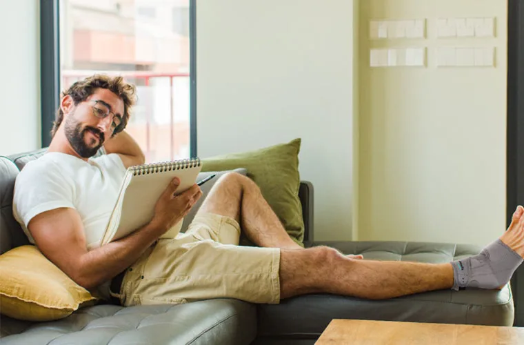 Man relaxing on a sofa with a notebook, wearing the grey Tynor Ankle Support (Neoprene)