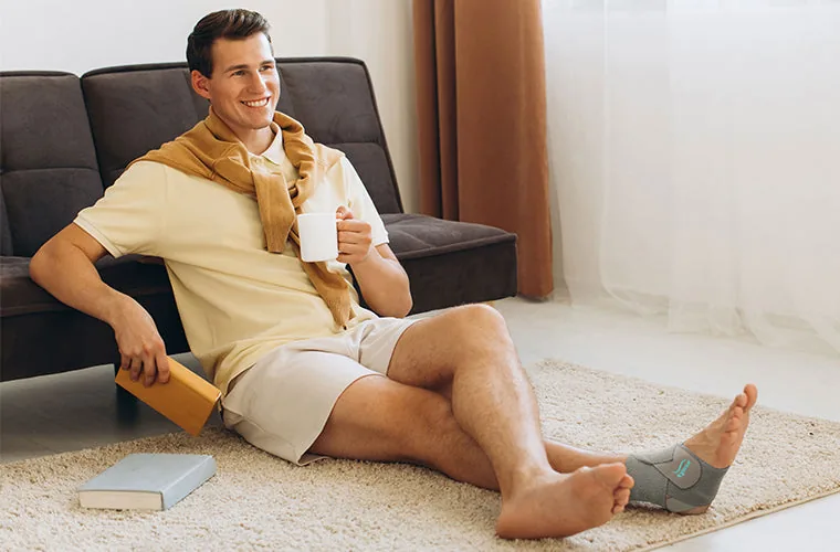 Man sitting on a rug with a mug, wearing the Tynor Ankle Wrap Sleek Elastic