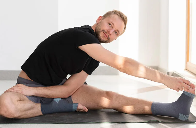 Man in a black T-shirt doing a seated stretch on a mat, wearing the Tynor Anklet Comfeel on both ankles