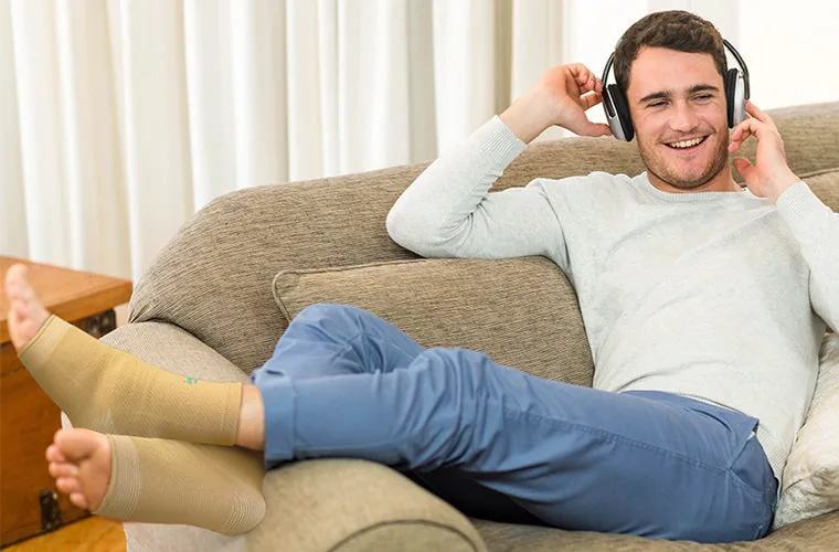 Smiling man in headphones lying back on a sofa with his feet up, wearing the Tynor Anklet on both feet