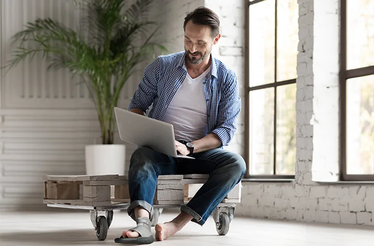 Man in a checked shirt using a laptop on a wooden pallet seat, wearing the Tynor Cast Shoe on one foot