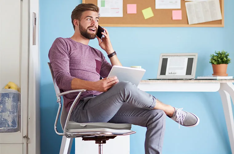 Smiling man on a phone call with a tablet in hand, sitting on the Tynor Coccyx Cushion Seat at a home desk