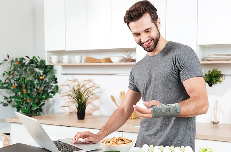 Smiling man in a grey T-shirt at a kitchen counter with a laptop, wearing the Tynor Elastic Wrist Splint