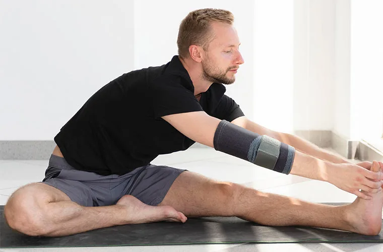 Man in a black T-shirt stretching forward on a yoga mat, wearing the Tynor Elbow Support