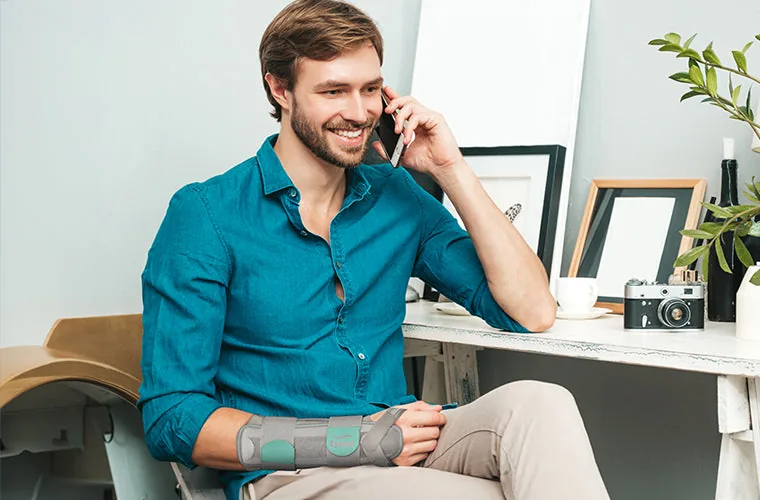 Smiling man in a teal shirt on a phone call at his desk, wearing the Tynor Forearm Splint