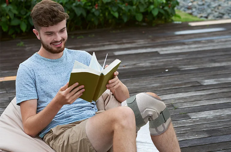 Young man reading a book outdoors on a bean bag, wearing the Tynor Functional Knee Support