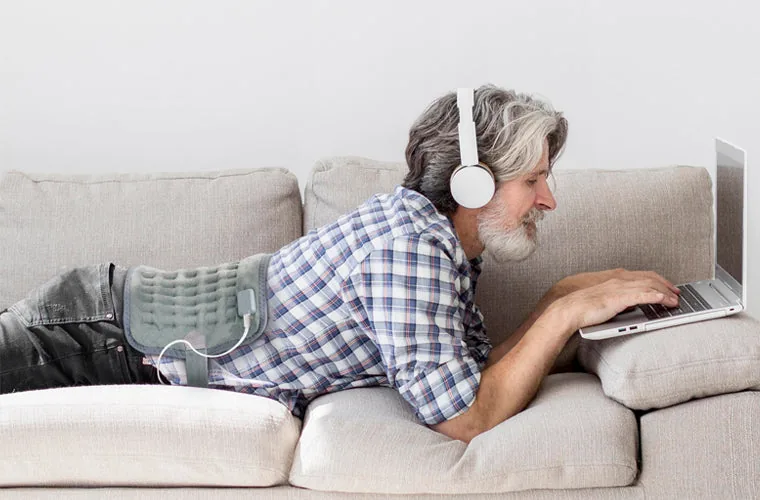 Grey-haired man in headphones lying on a sofa with a laptop, the Tynor Heating Pad Ortho on his lower back