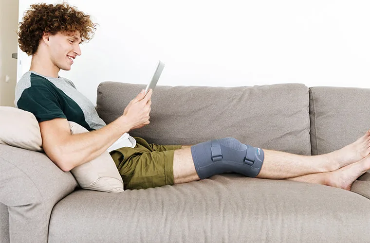 Young man with curly hair lying on a sofa reading a tablet, wearing the grey Tynor Knee Cap With Rigid Hinge