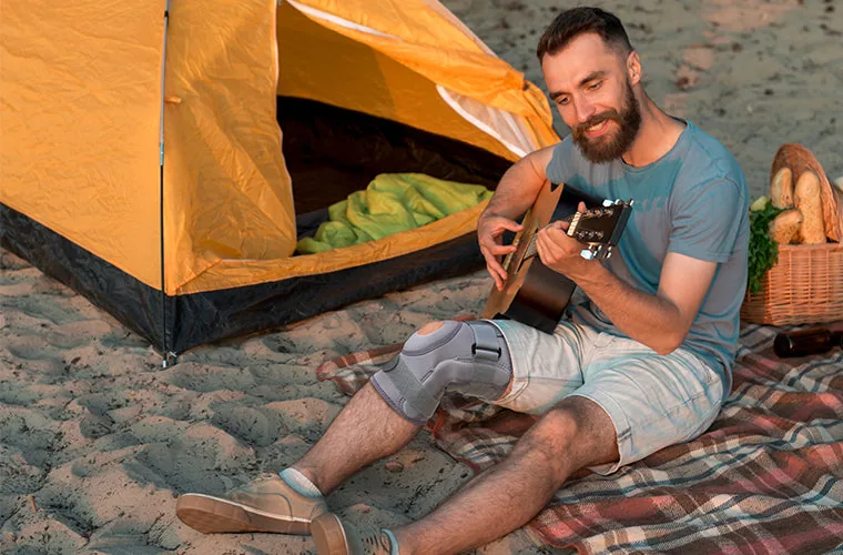 Man playing guitar on a picnic blanket beside a tent on the beach, wearing the Tynor Knee Support Hinged (Neoprene)