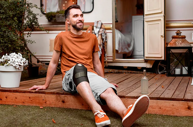 Smiling man in an orange T-shirt sitting on a wooden deck outside a caravan, wearing the Tynor Knee Wrap Hinged (Neoprene)