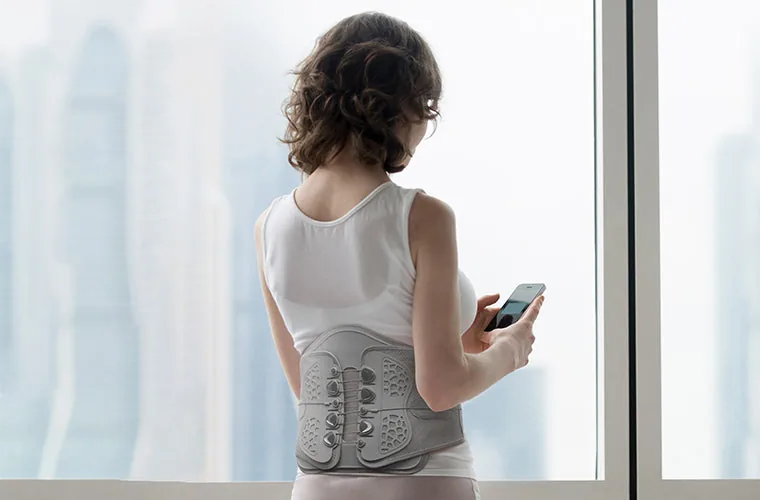 Woman in a white vest standing at a window with her phone, wearing the Tynor Lumbo Lacepull Brace