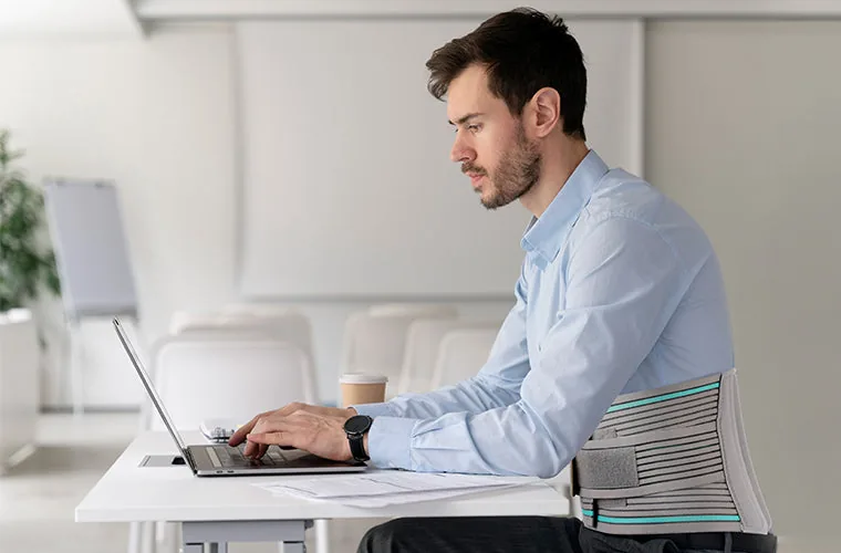 Man in a blue shirt working on a laptop at a white desk, wearing the Tynor Lumbo Sacral Belt