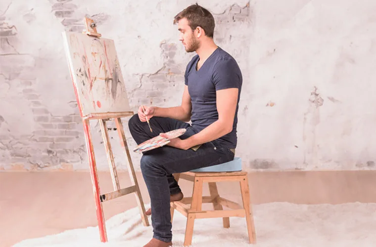 Man in a navy T-shirt painting at an easel while seated on the Tynor Ortho Cushion Seat on a wooden stool