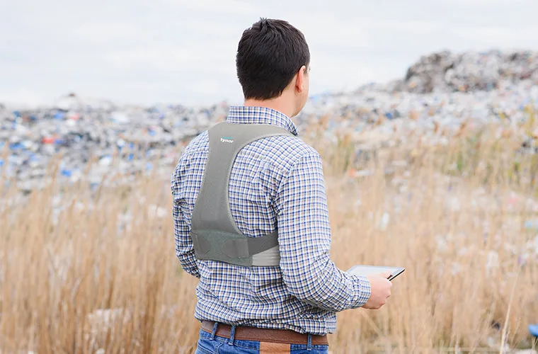 Man in a checked shirt holding a phone in a field, wearing the Tynor Posture Corrector, seen from behind