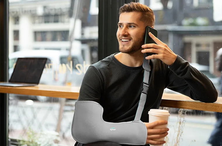 Smiling man in a black top on a phone call at a cafe table, holding a coffee cup, wearing the Tynor Pouch Arm Sling Tropical