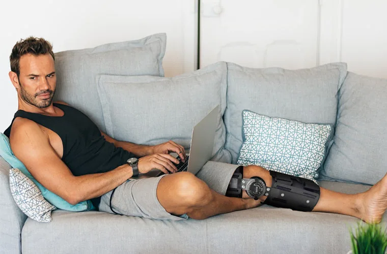 Man using a laptop on a sofa while wearing the Tynor R.O.M. Knee Brace 18 in