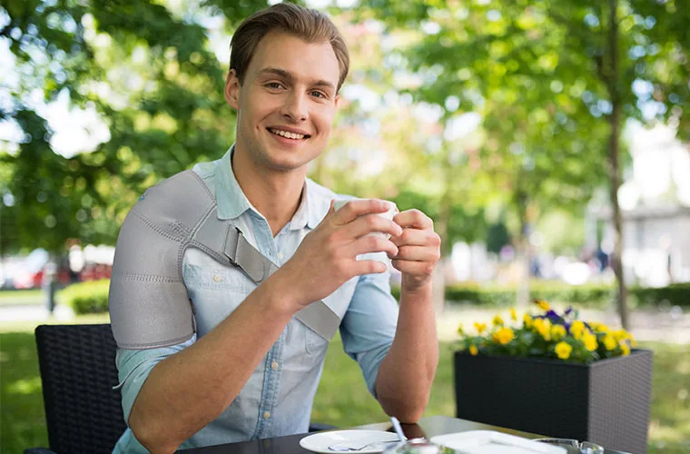 Smiling man in a light denim shirt at an outdoor cafe table, wearing the Tynor Shoulder Support (Neoprene)