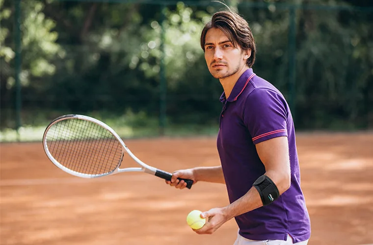 Man in a purple polo with a racket and ball on a tennis court, wearing the Tynor Tennis Elbow Support