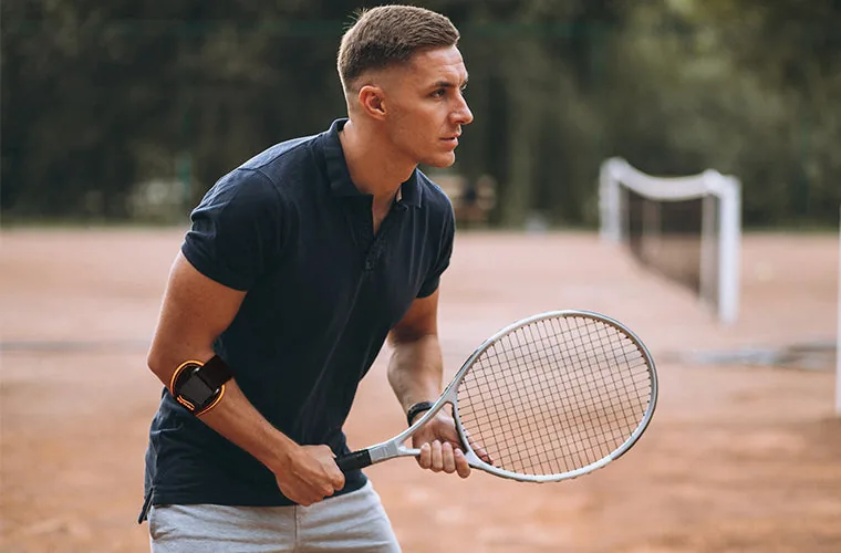 Man in a navy polo waiting to return a shot on a clay court, wearing the Tynor Tennis/Golfer's Elbow Support Pro