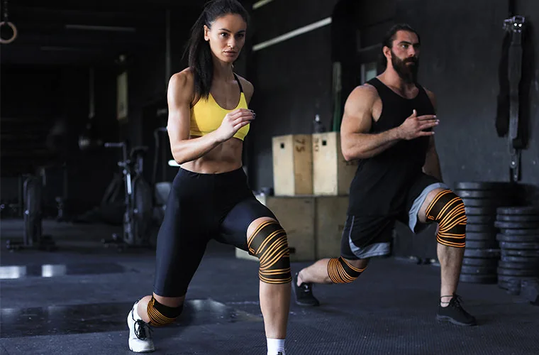 Woman and man doing lunges in a gym, both wearing the Tynor Weight Lifting Knee Wrap in black and orange