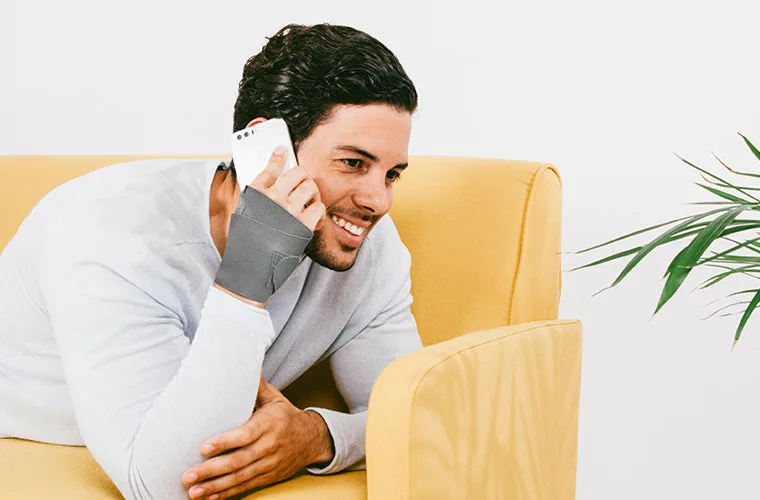 Man in a grey sweater talking on the phone by a yellow armchair, wearing the Tynor Wrist Brace With Thumb