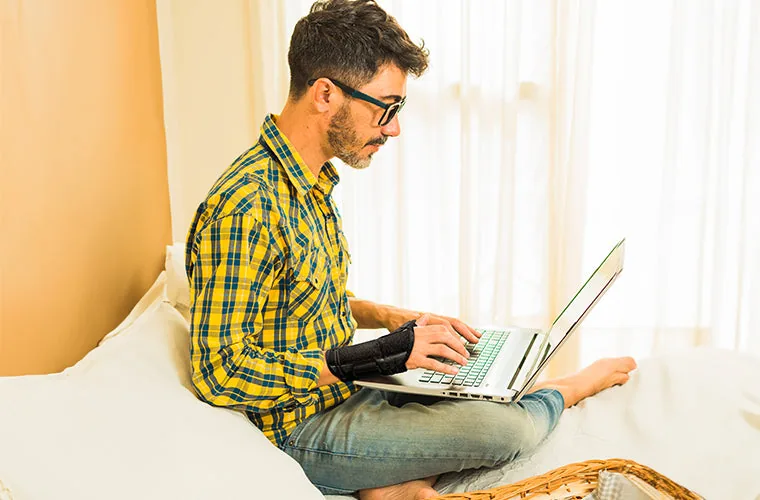 Man in a checked shirt sitting cross-legged on a bed with a laptop, wearing the Tynor Wrist Splint With Thumb